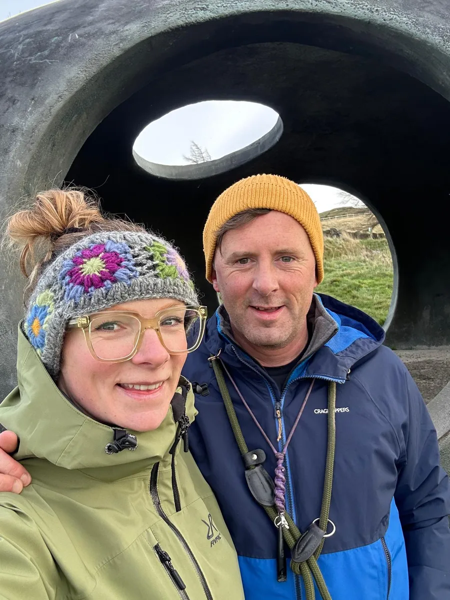 Nicola and Paul, the brother and sister who run Fettle It, smiling together outdoors in front of a large stone sculpture